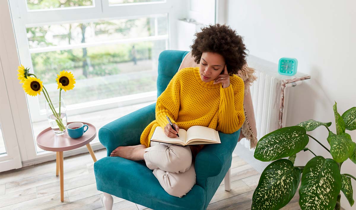Woman sitting in a teal armchair, writing in a notebook, enhancing her creative thinking in a serene and cozy environment.